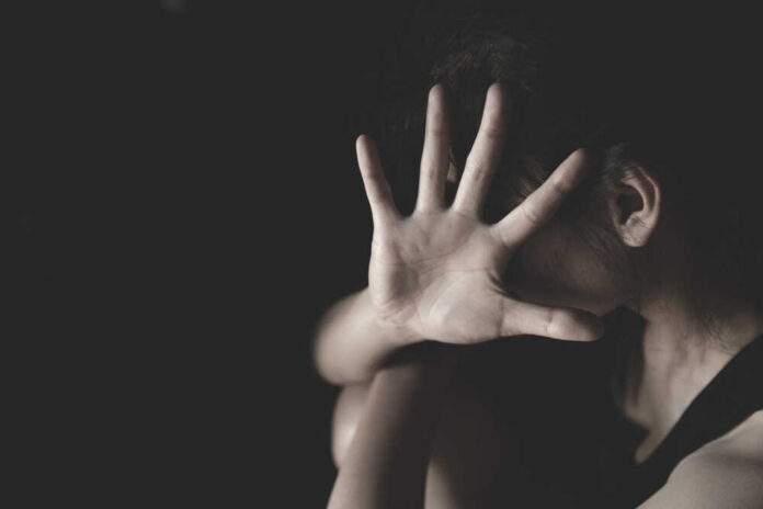 A person with their back turned, covering their face with a hand against a dark background