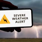 A hand holding a smartphone displaying a severe weather alert against a stormy background