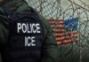 Police officer in tactical gear standing near a barbed wire fence with an American flag in the background