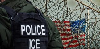 Police officer in tactical gear standing near a barbed wire fence with an American flag in the background