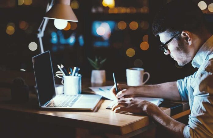 Person writing notes at a desk with a laptop and a warm light