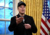 Man wearing hat gesturing in room with flag