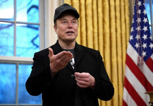 elon-musk2589898147jpg-1 Man wearing hat gesturing in room with flag