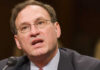 Man speaking into a microphone wearing glasses and suit