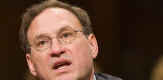 Man speaking into a microphone wearing glasses and suit