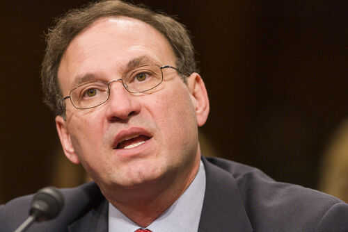 Man speaking into a microphone wearing glasses and suit