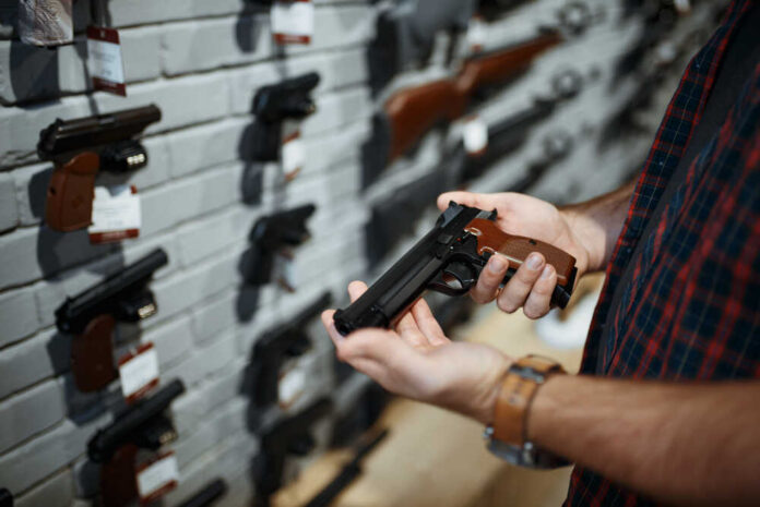 Man,Holds,Handgun,In,Gun,Shop Person holding a handgun in a gun shop with various firearms displayed on the wall