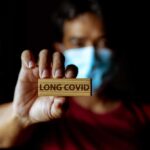 Person holding a wooden sign that reads 'LONG COVID' while wearing a mask