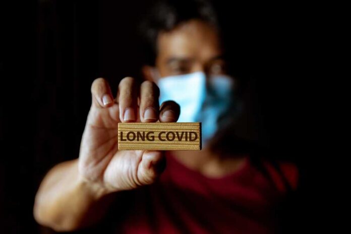 Person holding a wooden sign that reads 'LONG COVID' while wearing a mask