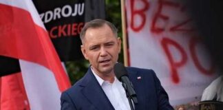 A speaker addressing a crowd at a political rally with flags in the background