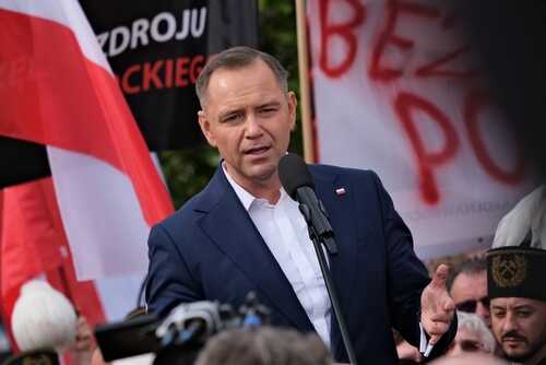 A speaker addressing a crowd at a political rally with flags in the background