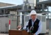 Man in a hard hat speaking at a podium in an industrial area