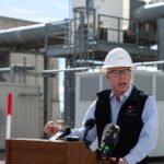 Man in a hard hat speaking at a podium in an industrial area