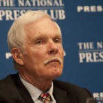 A man with gray hair and a mustache sitting at a press conference