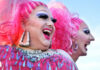 Two drag performers with bright pink hair and vibrant costumes laughing together