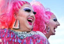 Two drag performers with bright pink hair and vibrant costumes laughing together