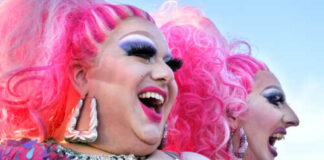 Two drag performers with bright pink hair and vibrant costumes laughing together