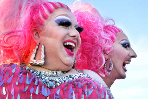 Two drag performers with bright pink hair and vibrant costumes laughing together