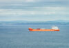 Hormuz Showdown: Who Really Controls It? Orange cargo ship sailing on calm sea with distant green hills