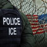 Questions Mount After ICE Custody Death Police officer in tactical gear standing near a barbed wire fence with an American flag in the background
