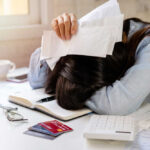 Economic Reality Isn’t Matching Official Numbers Woman with head on desk amid bills and credit cards