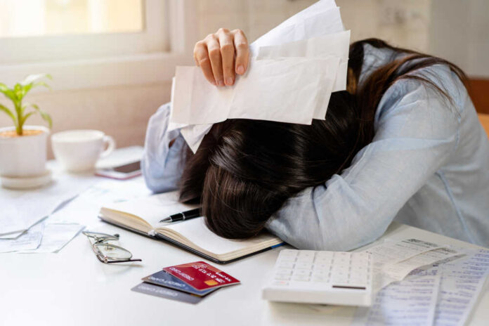 Stressed,Young,Woman,Calculating,Monthly,Home,Expenses,,Taxes,,Bank,Account Woman with head on desk amid bills and credit cards