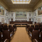 Impeachment Move, Sudden Death — What Happened? Interior view of a courtroom with wooden benches and large murals