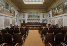 Impeachment Move, Sudden Death — What Happened? Interior view of a courtroom with wooden benches and large murals