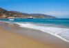 Sandy beach with gentle waves and a long pier by coastal hills