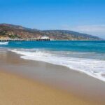 Heroic Beach Rescue Comes At Devastating Cost Sandy beach with gentle waves and a long pier by coastal hills