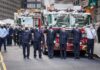 Firefighters in dress uniforms salute in front of fire trucks