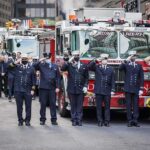 Twenty-Five Years Later, Their Courage Still Inspires Firefighters in dress uniforms salute in front of fire trucks