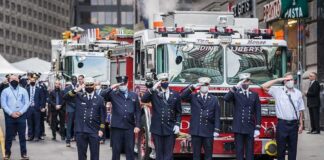 Firefighters in dress uniforms salute in front of fire trucks