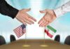 War Pause Sparks Fury—Who’s Lying Here? Two hands reaching to shake above US and Iran flags on a conference table