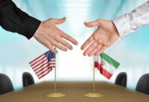 Two hands reaching to shake above US and Iran flags on a conference table