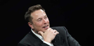 Elon Musk speaking on stage in a dark suit, resting his chin on his hand