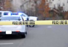 Police tape cordons off a street with patrol cars and flashing lights in the background