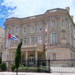Cuba Cries Blockade – Talks Denied neoclassical stone building with a Cuban flag and iron fence