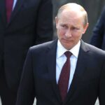 Vladimir Putin in a dark suit with a red tie walking outdoors