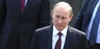 Vladimir Putin in a dark suit with a red tie walking outdoors
