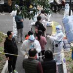 WHO’s Ebola FAILURE – Who’s to Blame? Christian Jones and others wait at an outdoor COVID-19 testing site with staff in protective suits