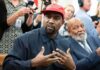 Kanye West speaking while Jim Brown sits nearby in a crowded room