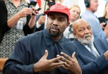 Kanye West speaking while Jim Brown sits nearby in a crowded room