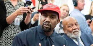 Kanye West speaking while Jim Brown sits nearby in a crowded room