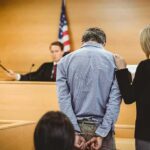 Judge Rejects Bid To Keep Witness Detained Defendant in handcuffs stands before a judge in a courtroom