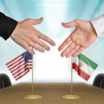 Tehran Says It Will Move—After America Does Two hands reaching to shake above US and Iran flags on a conference table