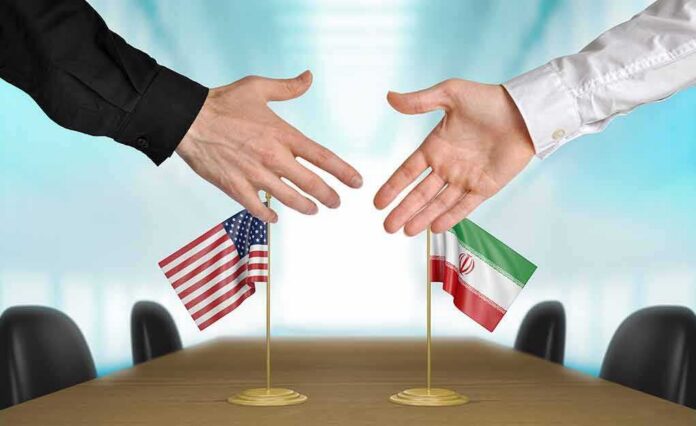 Two hands reaching to shake above US and Iran flags on a conference table