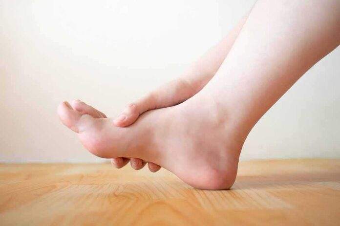 shutterstock_1186138735 Hand holding the arch of a bare foot on a wooden floor