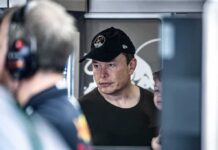 Elon Musk wearing a black cap in a crowded indoor setting