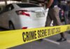 Yellow crime scene tape in front of a white car with investigators nearby